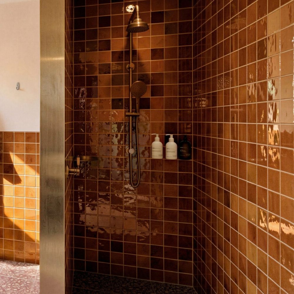 A walk-in shower with glossy brown tile, a bronze shower fixture, toiletries on a ledge, and a green robe hanging nearby.