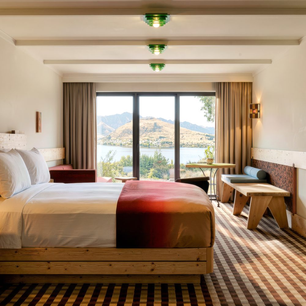 A wood-furnished hotel room with a large bed, seating area, and wide windows overlooking a lake and mountains.
