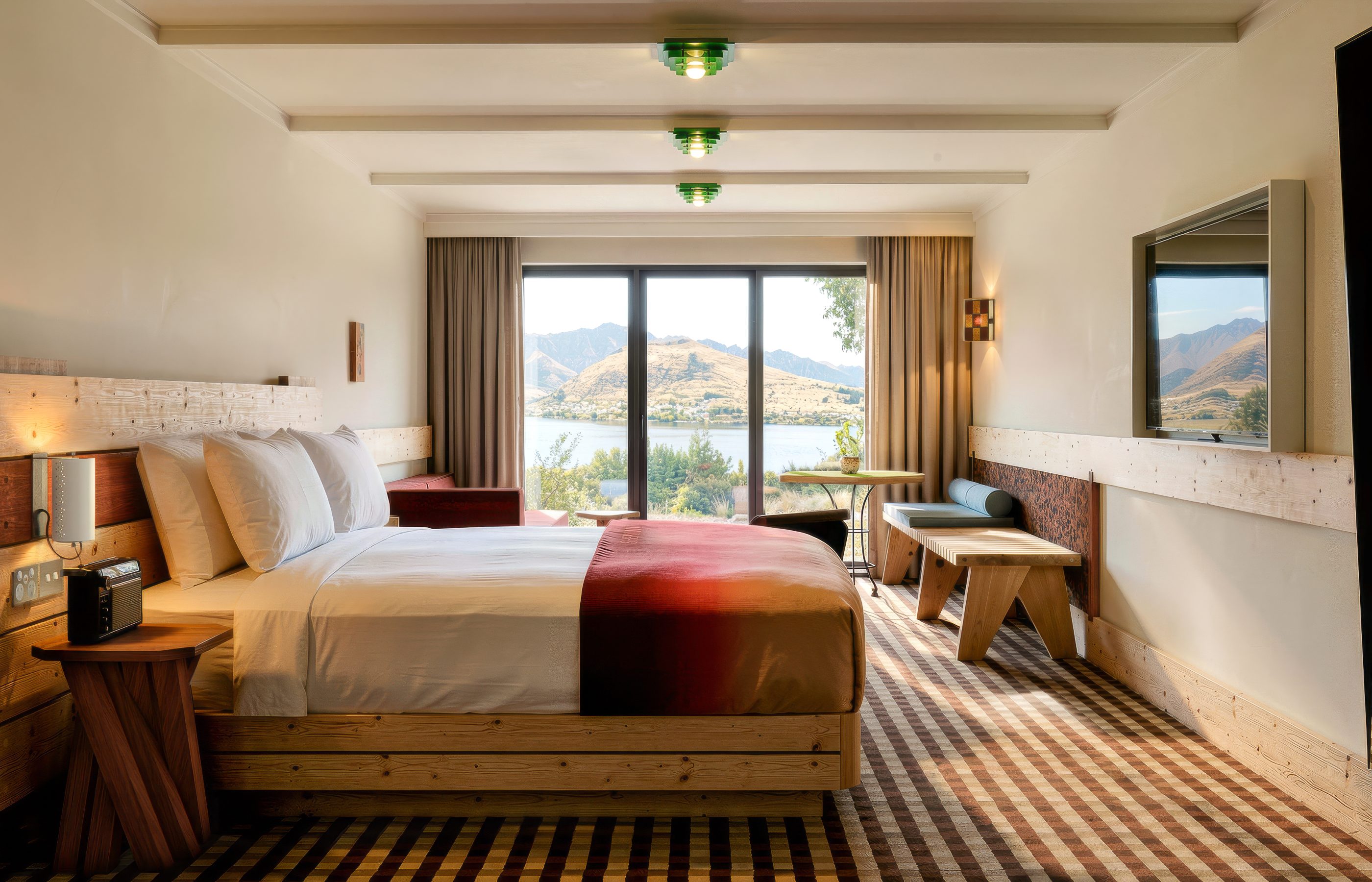 A wood-furnished hotel room with a large bed, seating area, and wide windows overlooking a lake and mountains.
