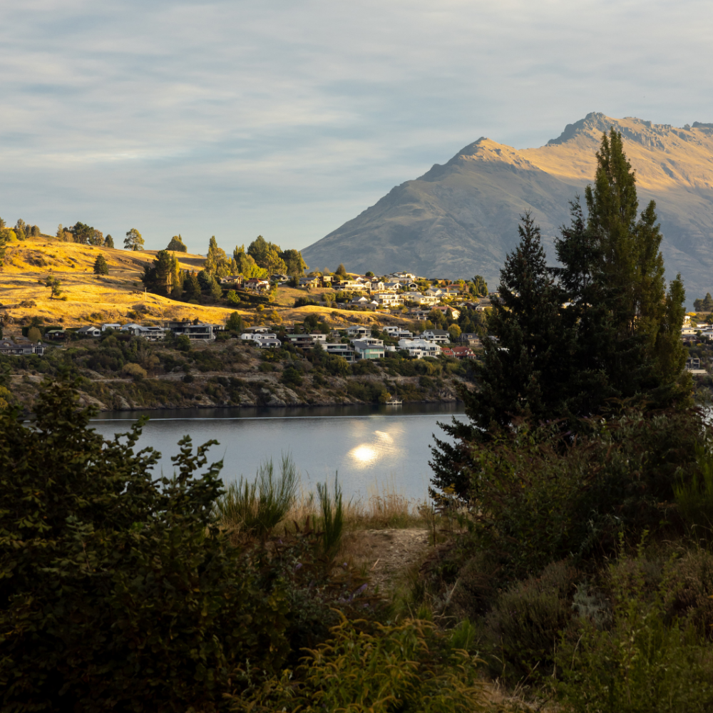 Sunlight illuminates a mountain above a lakeside town, with trees and shrubs framing the calm water.