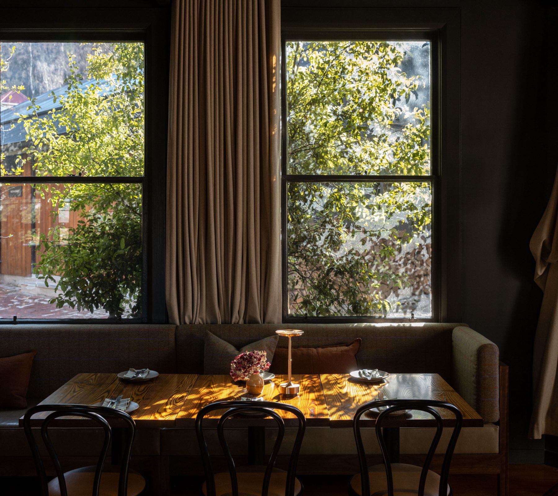 Sunlight falls across set wooden tables and a cushioned booth beneath tall curtained windows overlooking leafy trees.