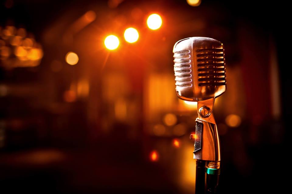 A vintage silver microphone stands on a dim stage, lit by glowing orange spotlights and blurred background lights.