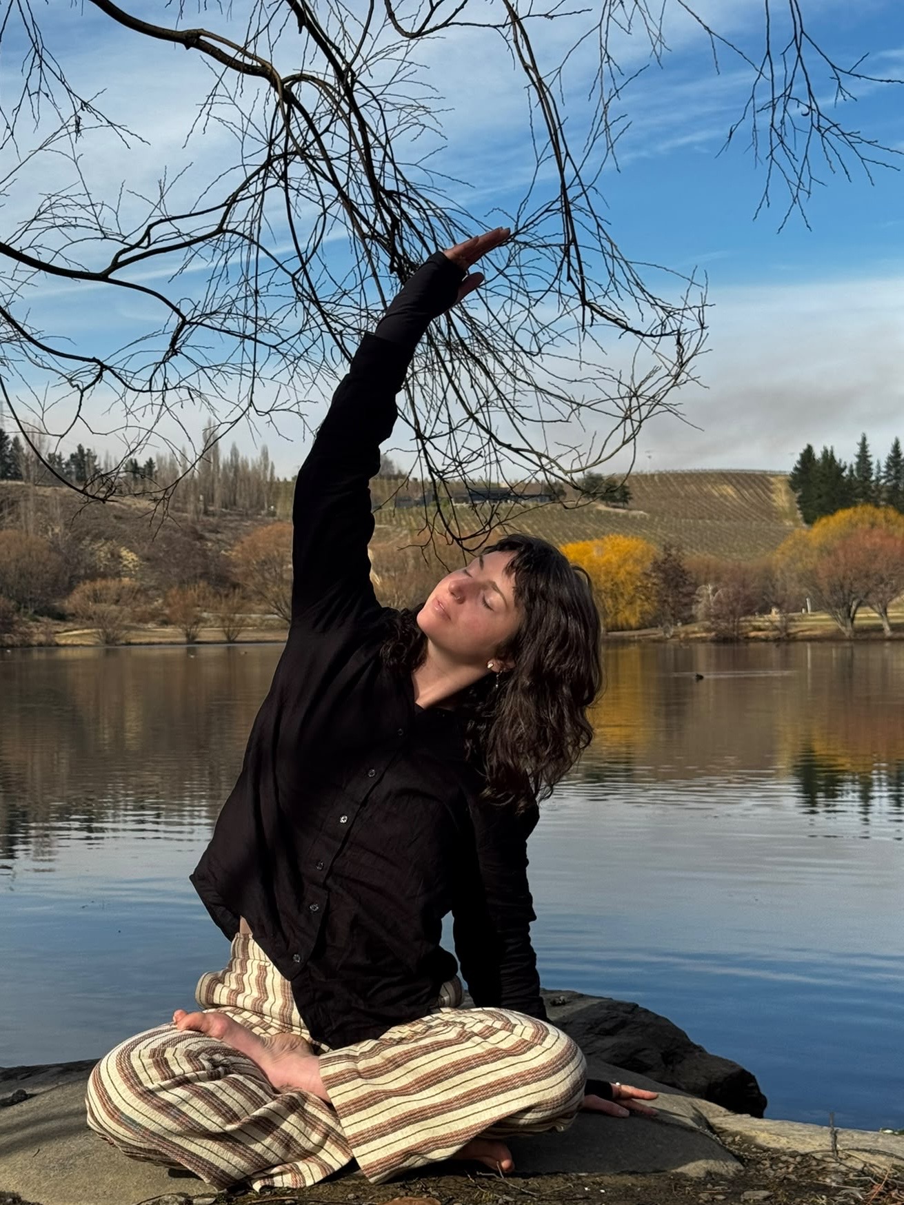 A woman sits cross-legged on rocks beside a calm lake, stretching one arm toward bare branches overhead.