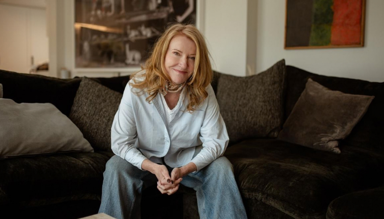 A red-haired woman in a light blue shirt and jeans sits on a dark sofa, hands clasped, with cushions and framed art behind her.
