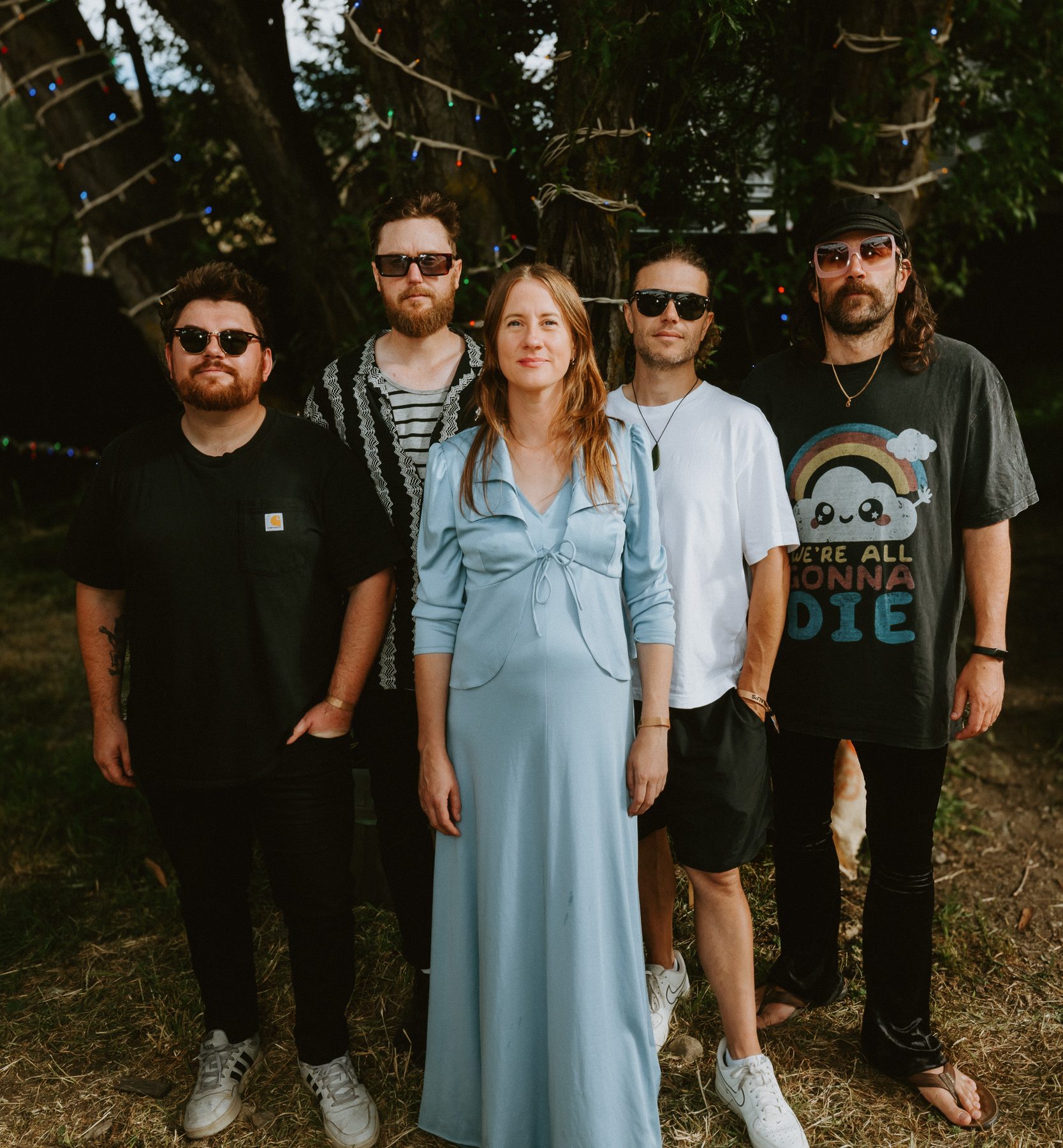 Five adults pose beneath trees strung with small colored lights; a woman in a blue dress stands between four men.
