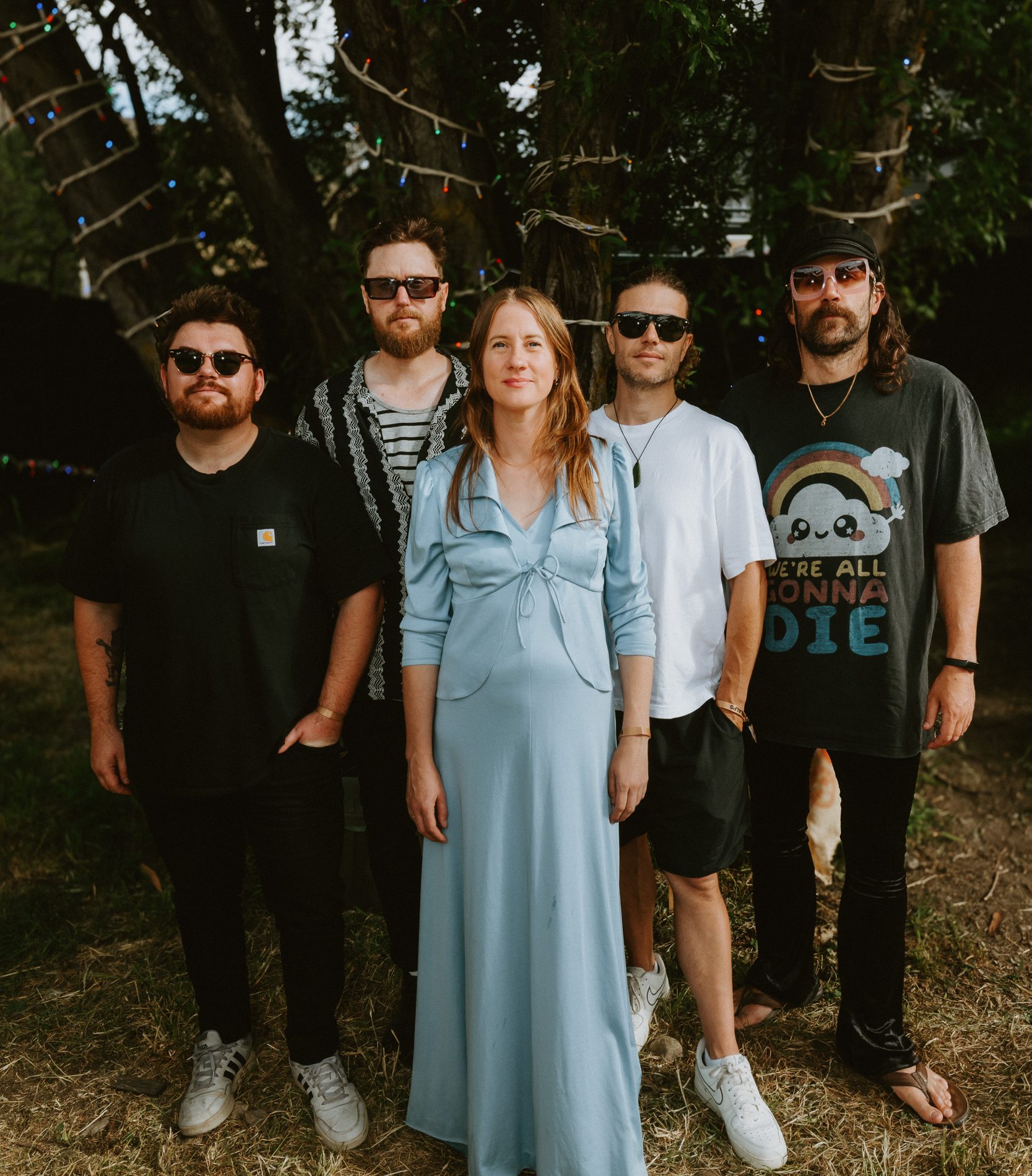 Five adults pose beneath trees strung with small colored lights; a woman in a blue dress stands between four men.