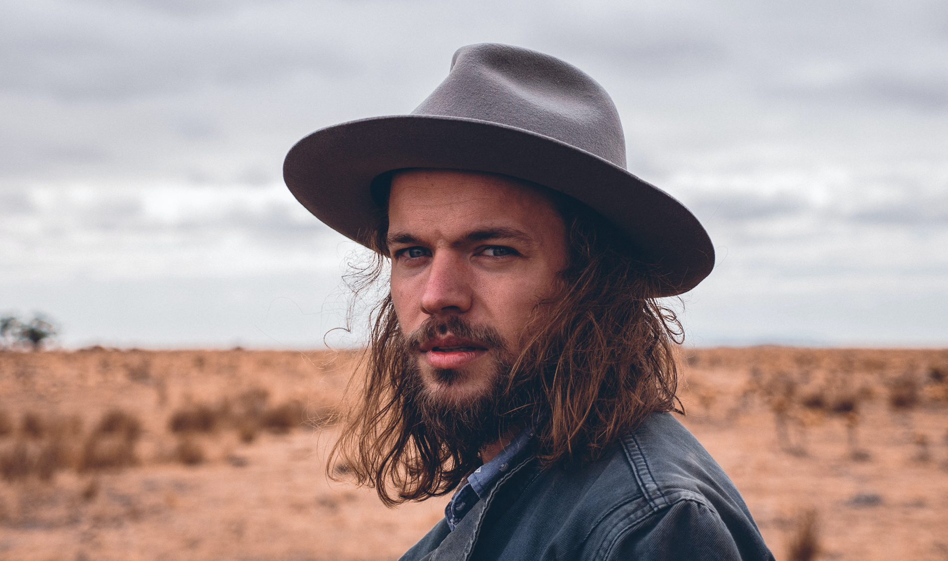 Close-up of a long-haired, bearded man in a wide-brimmed hat standing in a dry, open landscape.