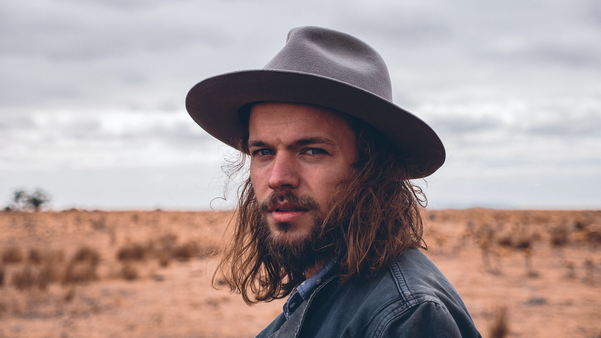 Close-up of a long-haired, bearded man in a wide-brimmed hat standing in a dry, open landscape.