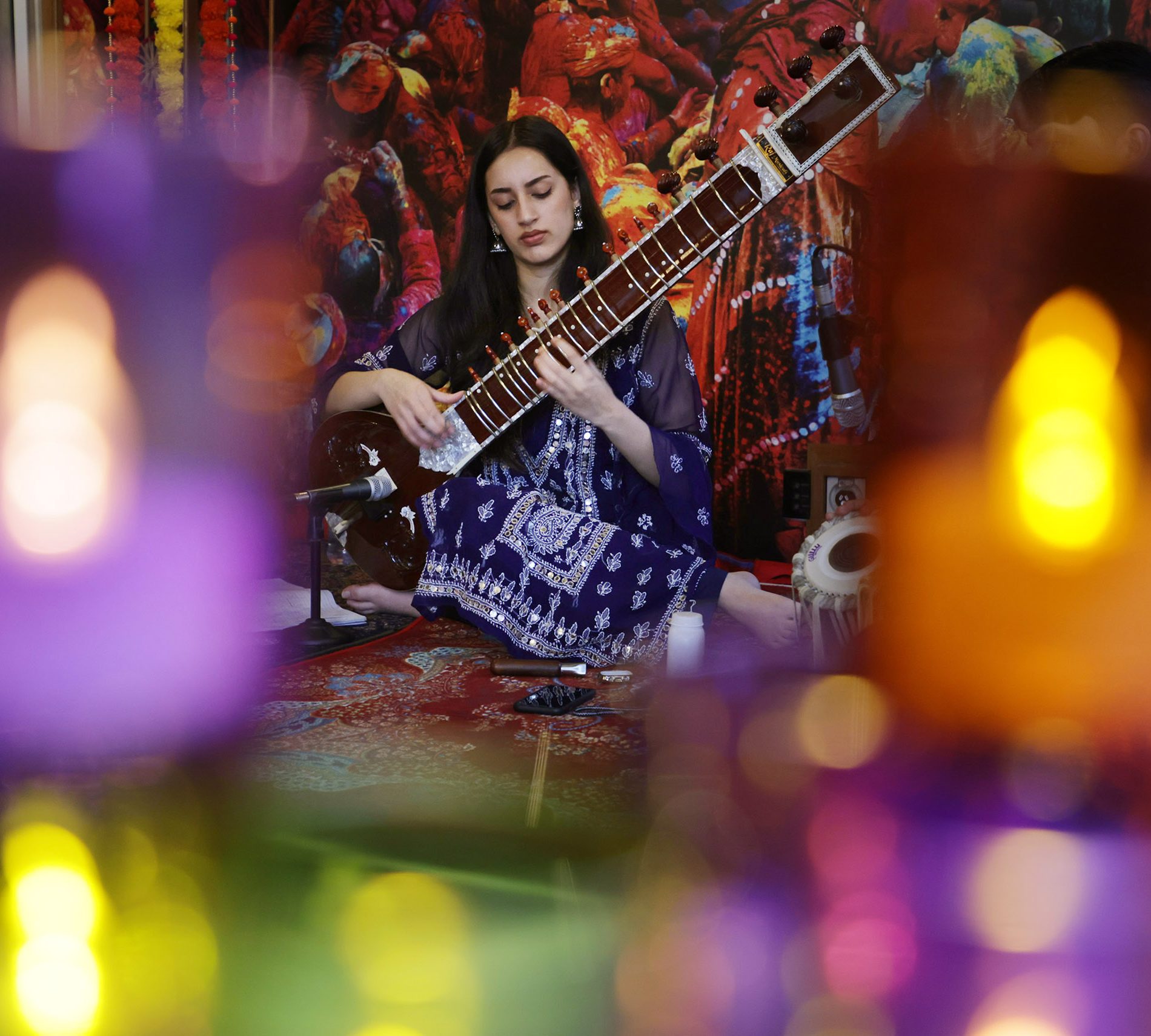 A seated woman plays a sitar before a colorful mural, with blurred multicolored lights in the foreground.